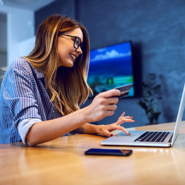 Side view of young charming caucasian woman with eyeglasses holding credit card and using laptop for online shopping while sitting at dining table at home.